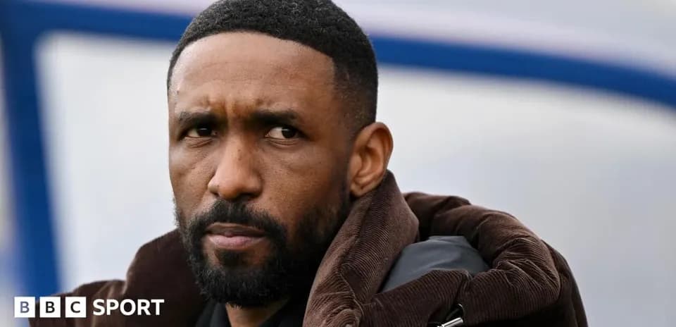 Man in a brown coat observing from sidelines