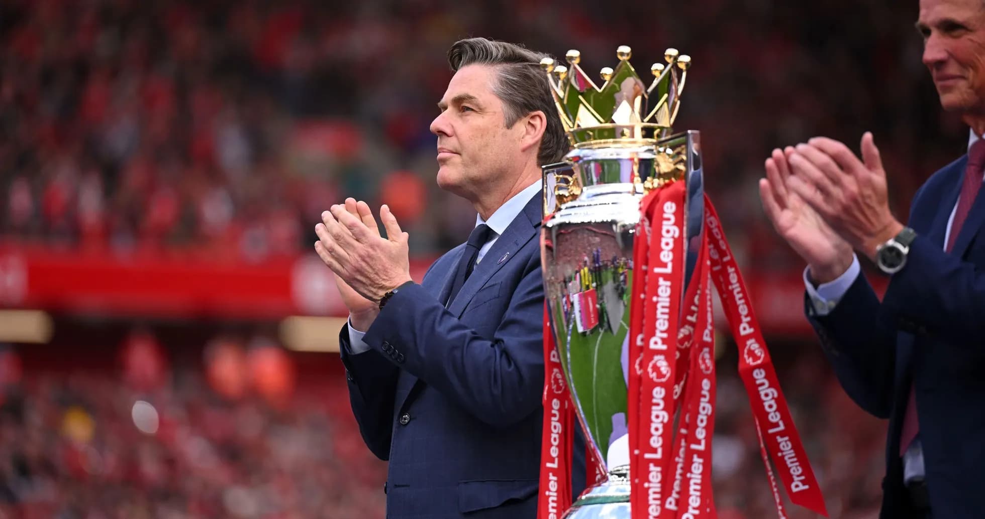 Man in suit applauding near Premier League trophy